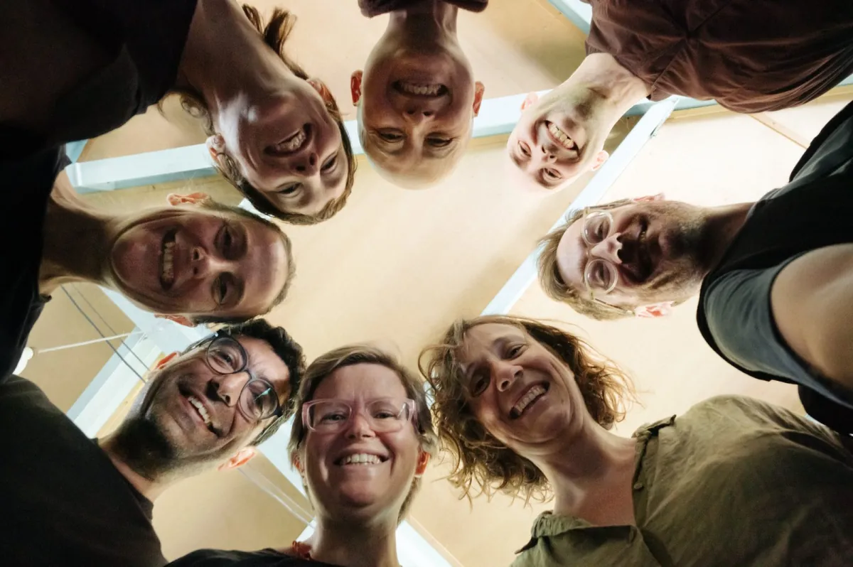 Eight friends in a circle with their heads together, seen from below, all smiling down at the camera.