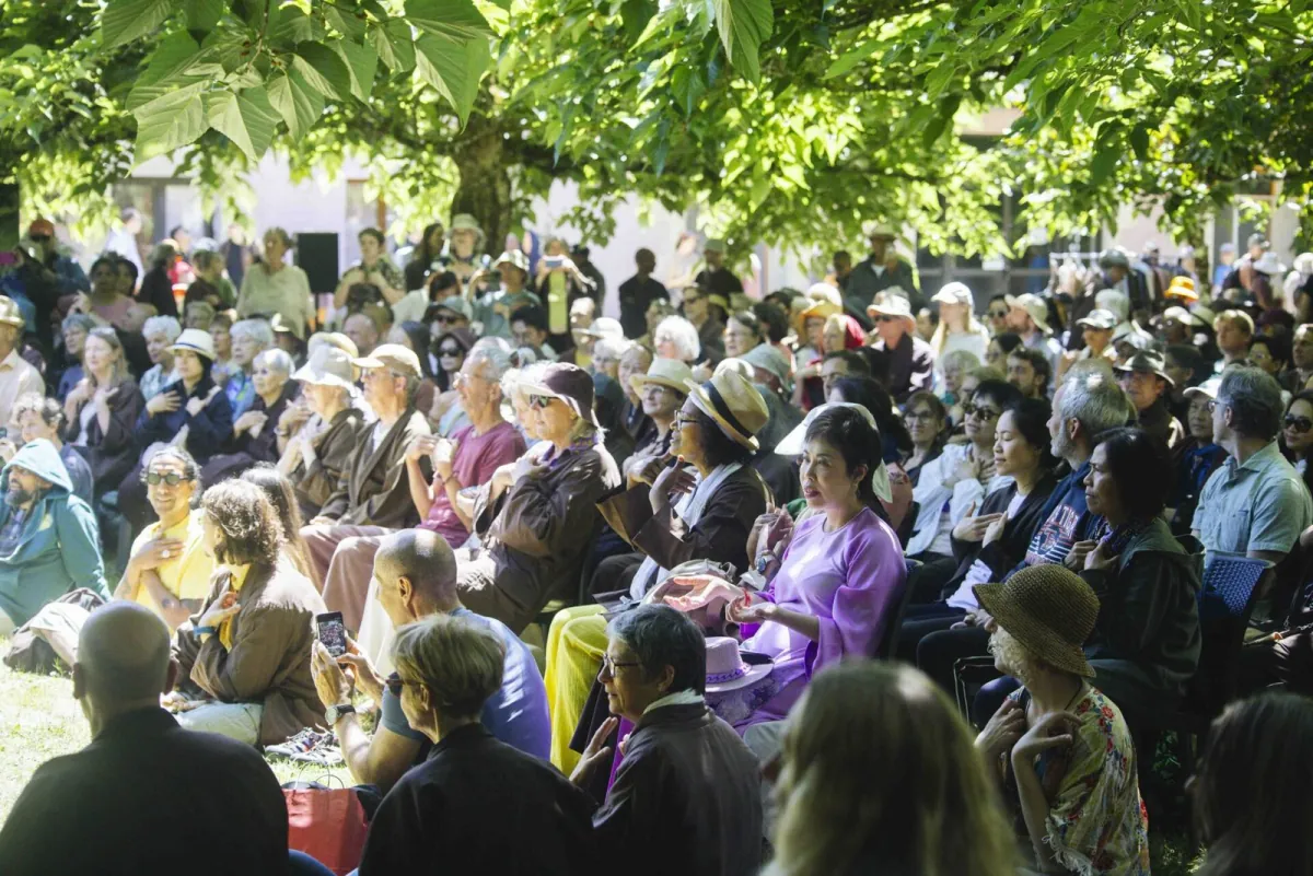 A crowd of friends seated under the trees in dappled sunlight, many with a hand on their heart, listening together.