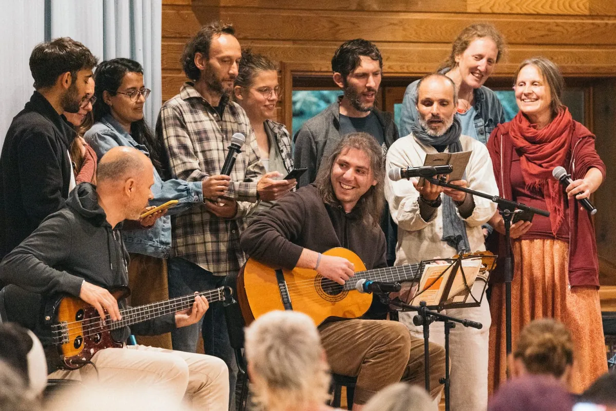 Friends making music together in a wooden hall, two of them playing guitars, others singing around them, with the backs of the listeners' heads in the foreground.