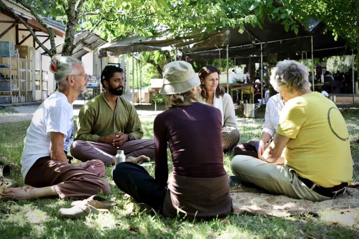 Six friends sitting in a circle on the grass in the shade of a tree, listening to one another, with a long tent and tables behind them.